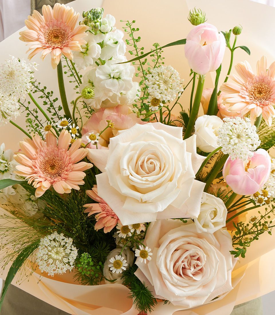 A pastel bouquet with cream roses, peach gerberas, pink tulips, white queen anne’s lace, and chamomile, wrapped in peach paper and tied with a satin ribbon.