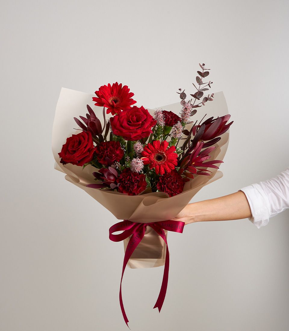 Elegant bouquet of red roses, gerberas, carnations, and foliage wrapped in beige paper with a red satin ribbon.