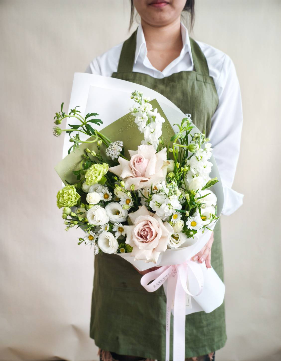 Elegant bouquet of pastel roses, white lisianthus, green carnations, and chamomile flowers, wrapped in white and green paper.
