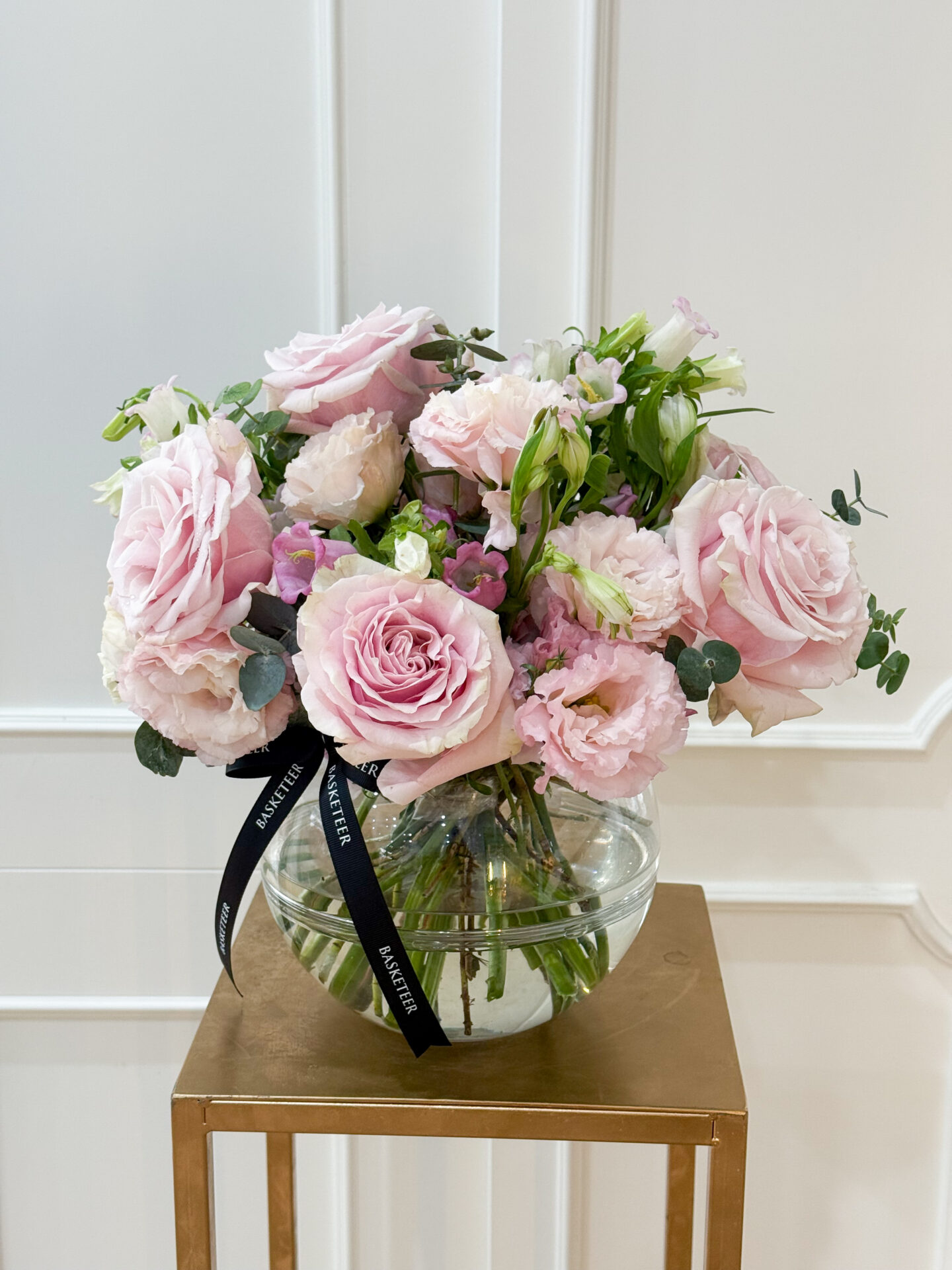Luxury glass vase filled with blush pink roses, lisianthus, spray roses, and soft greenery, tied with Basketeer ribbon.