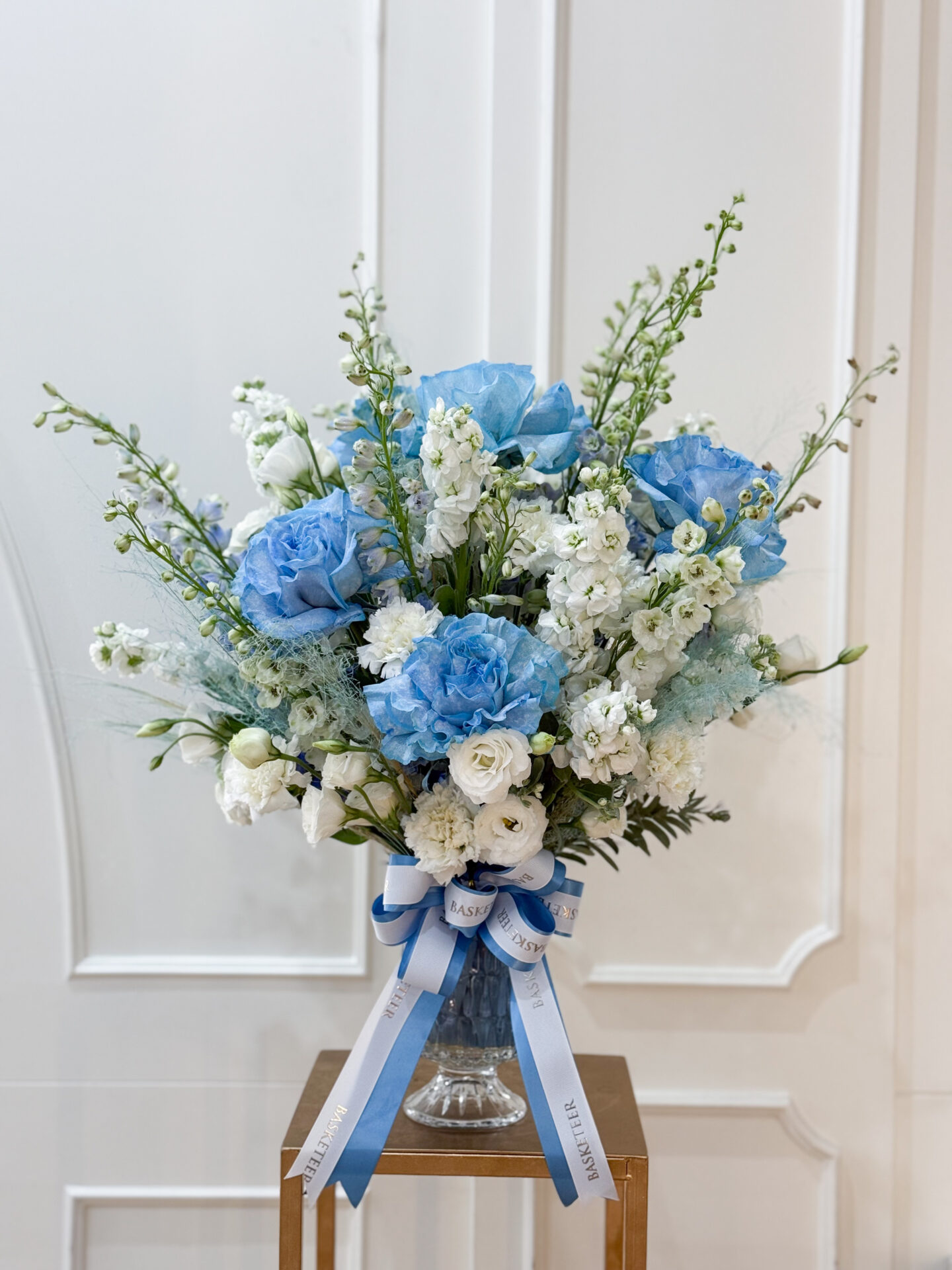 Luxury glass vase arrangement with blue roses, blue hydrangeas, white lisianthus, delphiniums, and lush greenery with ribbon bow.