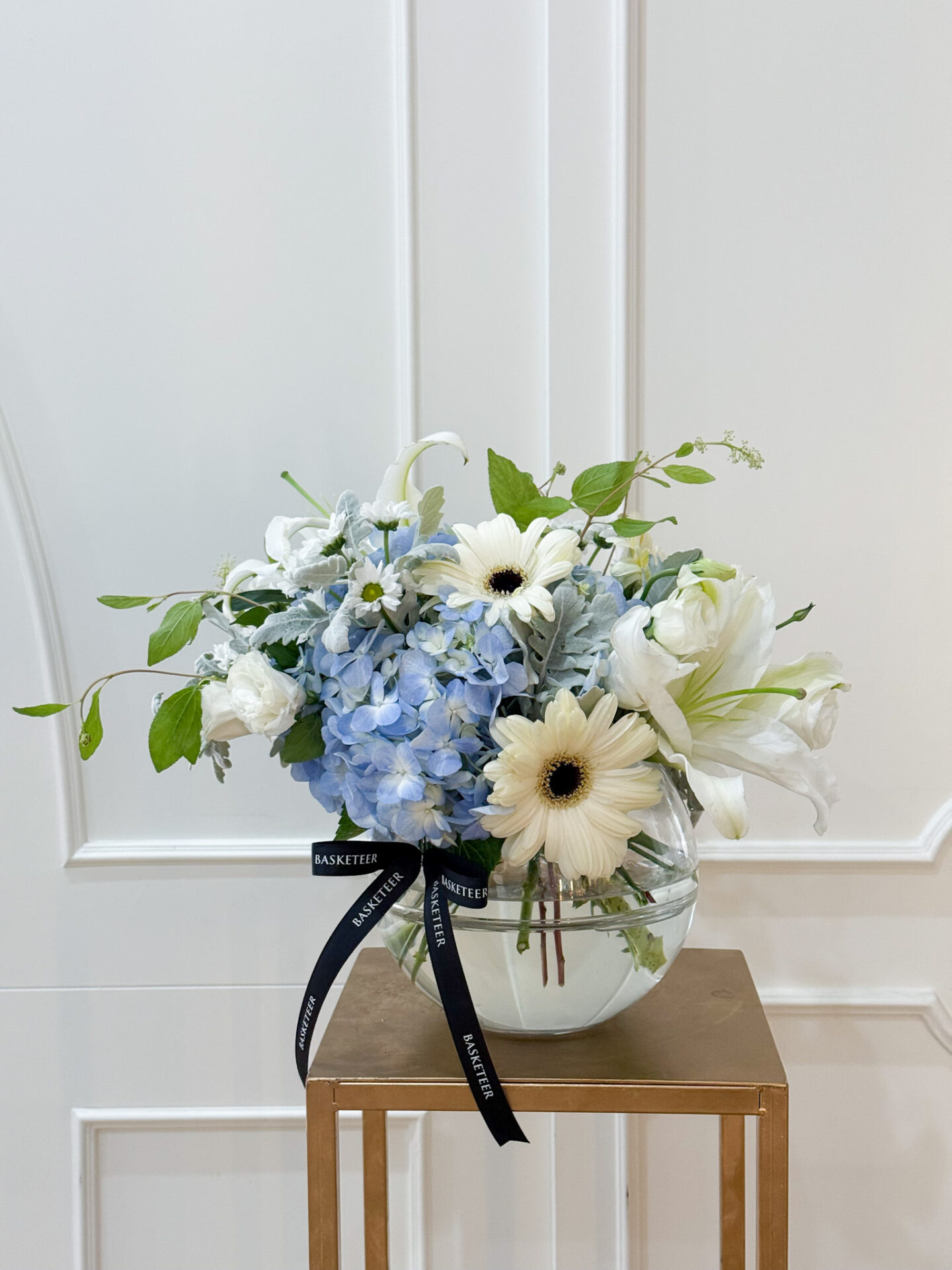 Elegant glass vase with blue hydrangeas, white lilies, gerberas, and fresh green leaves arranged by Basketeer