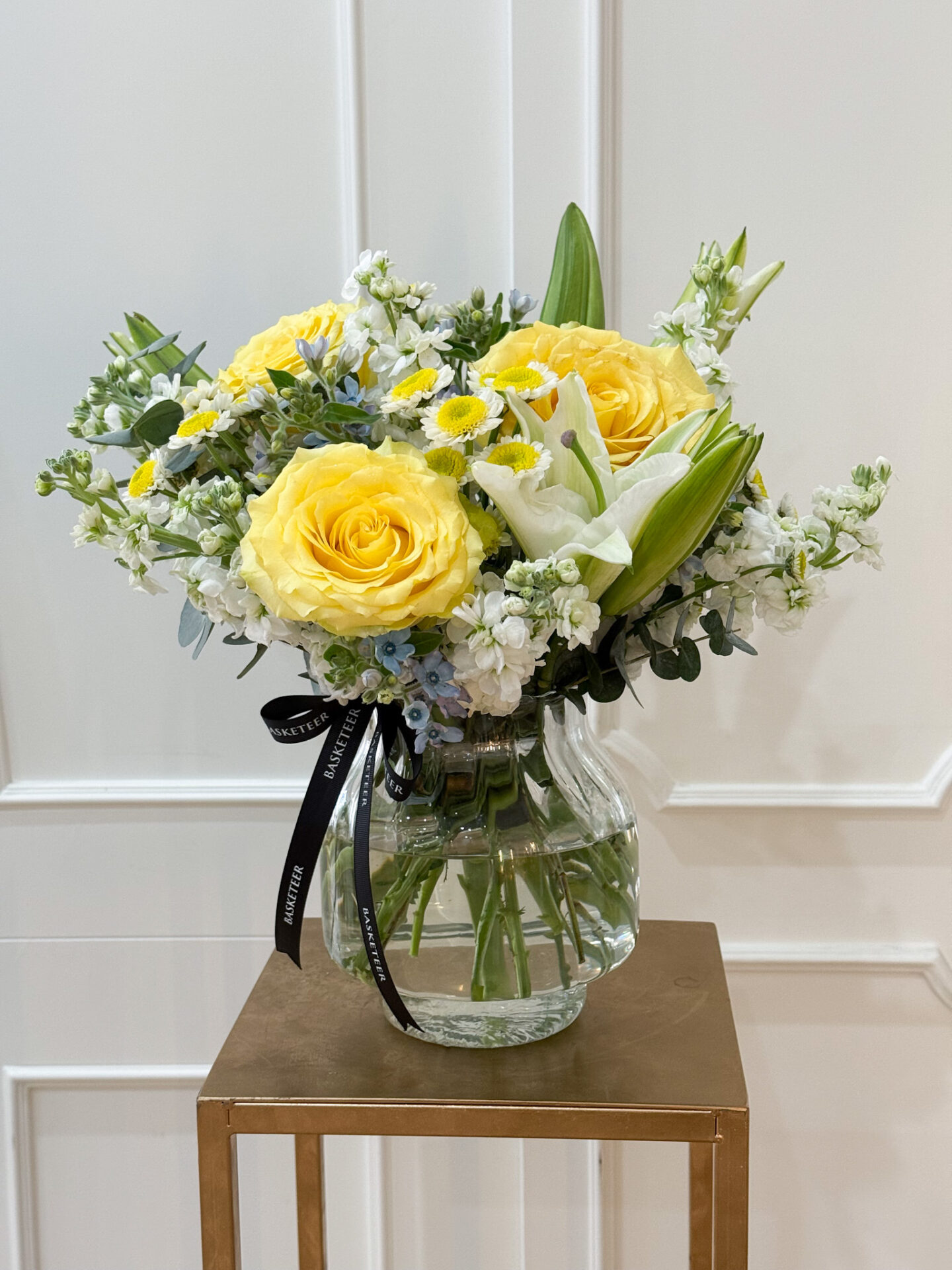 Elegant glass vase filled with yellow roses, white lilies, chamomile, and fresh greenery arranged by Basketeer