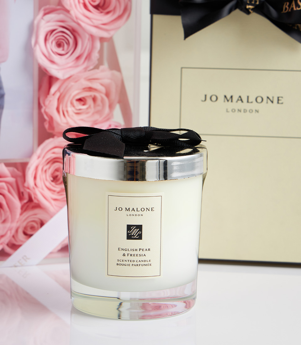 Close-up of Jo Malone London English Pear & Freesia scented candle with black bow, pink rose frame, and Jo Malone gift box in the background.