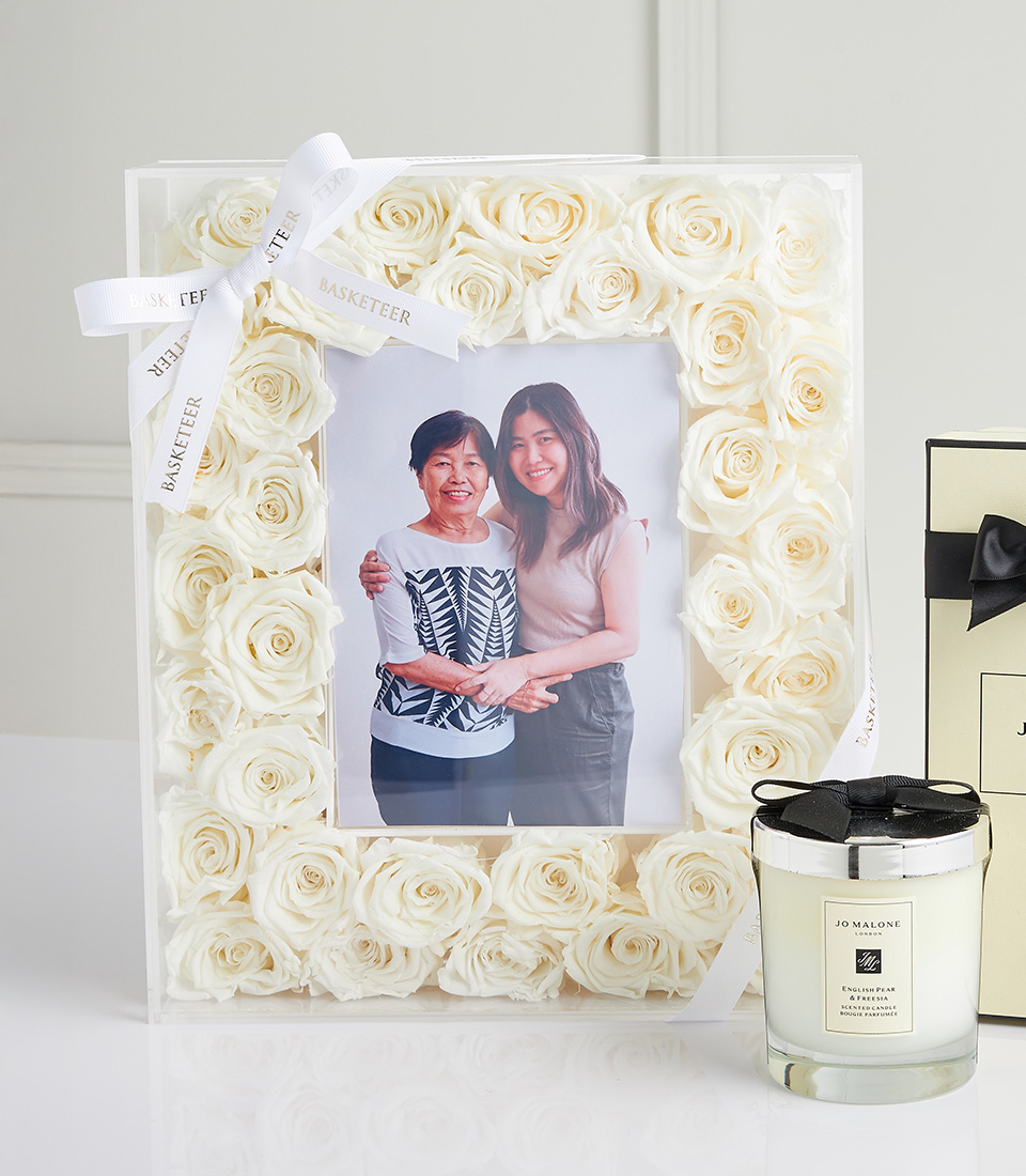 Close-up of a white rose photo frame with a mother and daughter picture, tied with a white ribbon, next to a Jo Malone scented candle.