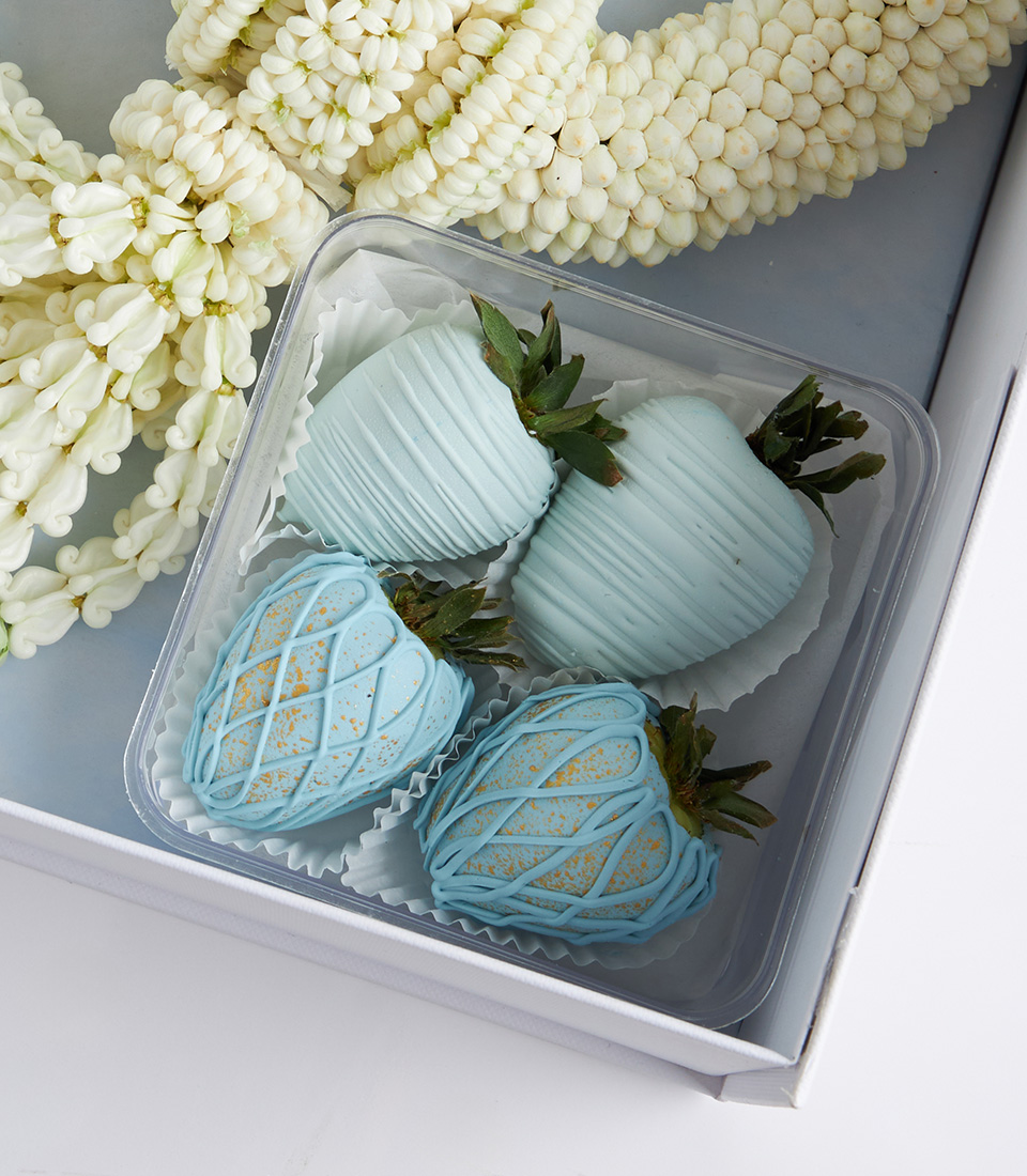 Close-up of pastel blue chocolate-covered strawberries in a gift box with a fresh jasmine garland.