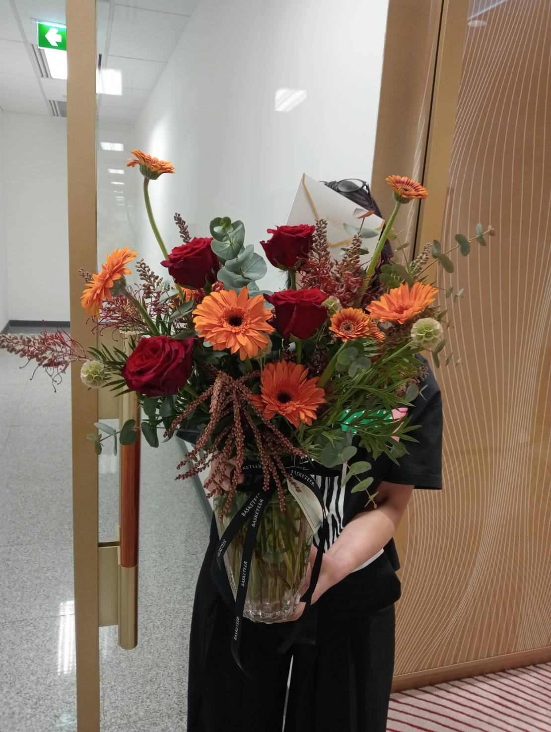 A person holding a Radiant Rouge Vase Arrangement filled with red roses, orange gerbera daisies, eucalyptus, and greenery stands indoors near a glass door, with a striped carpet visible on the floor.
