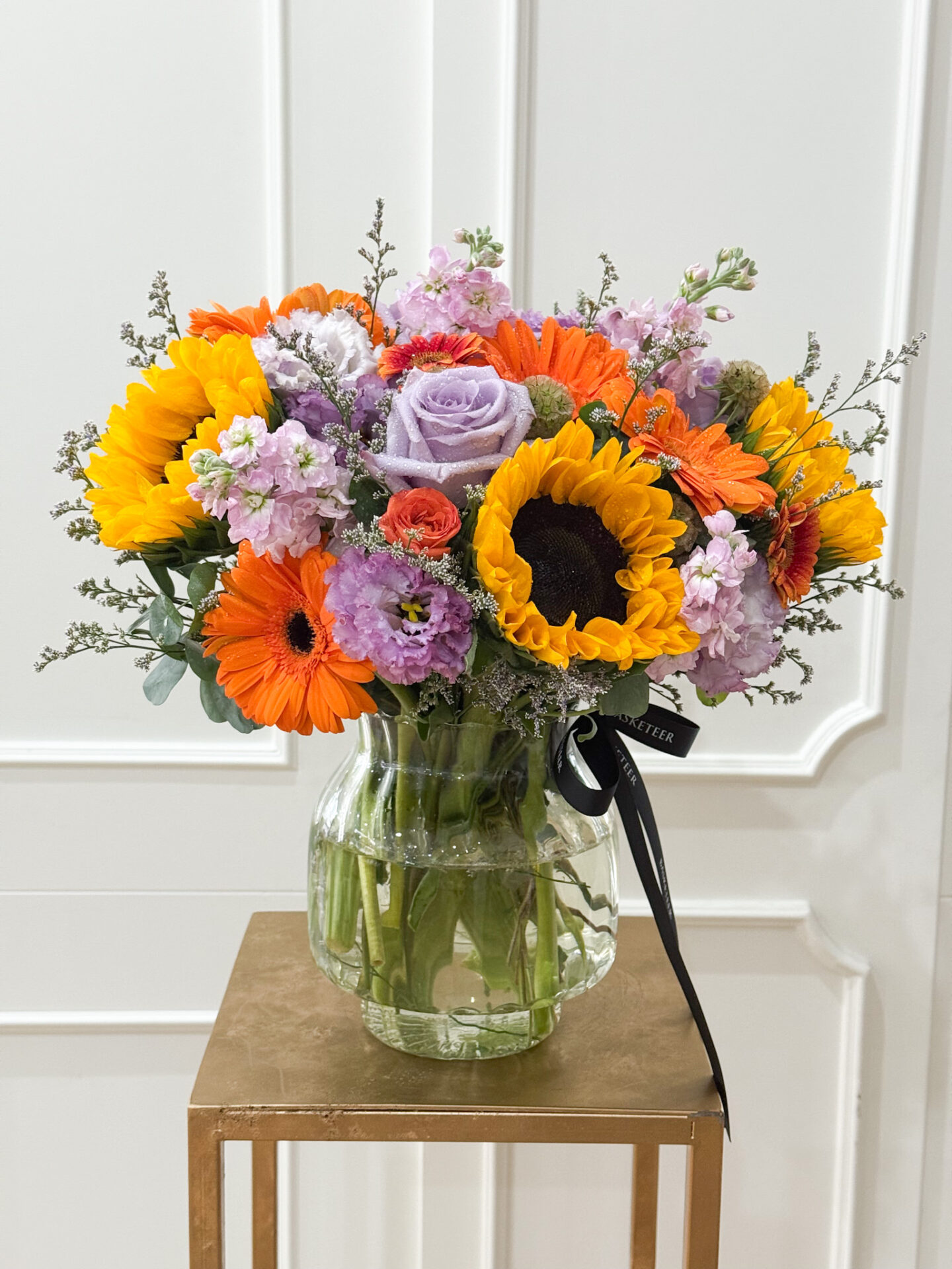 Bright vase arrangement with sunflowers, lavender roses, gerberas, and lisianthus, tied with Basketeer ribbon.