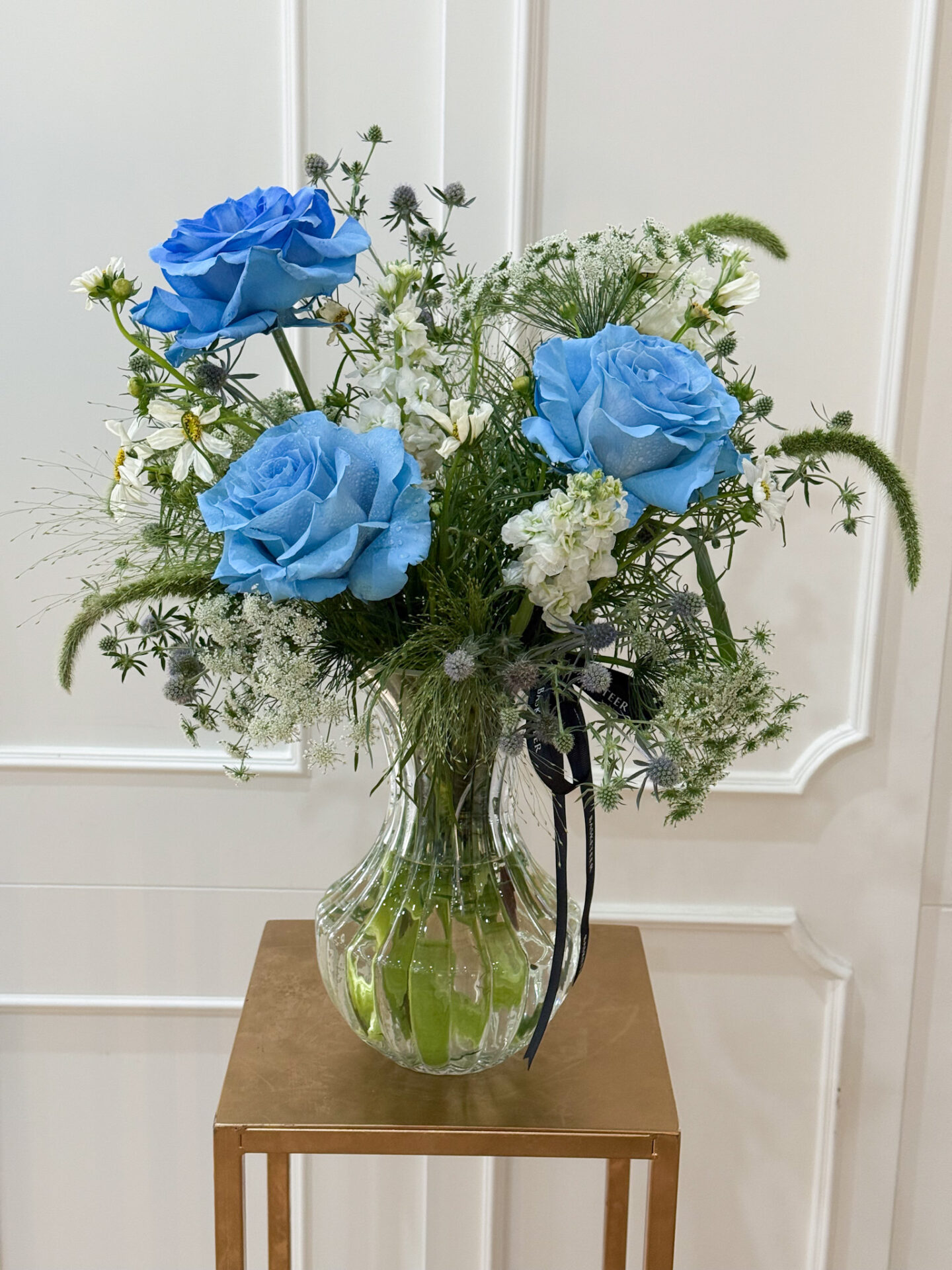 Elegant vase with rare blue roses, white filler flowers, and lush greenery arranged in a clear glass vase.