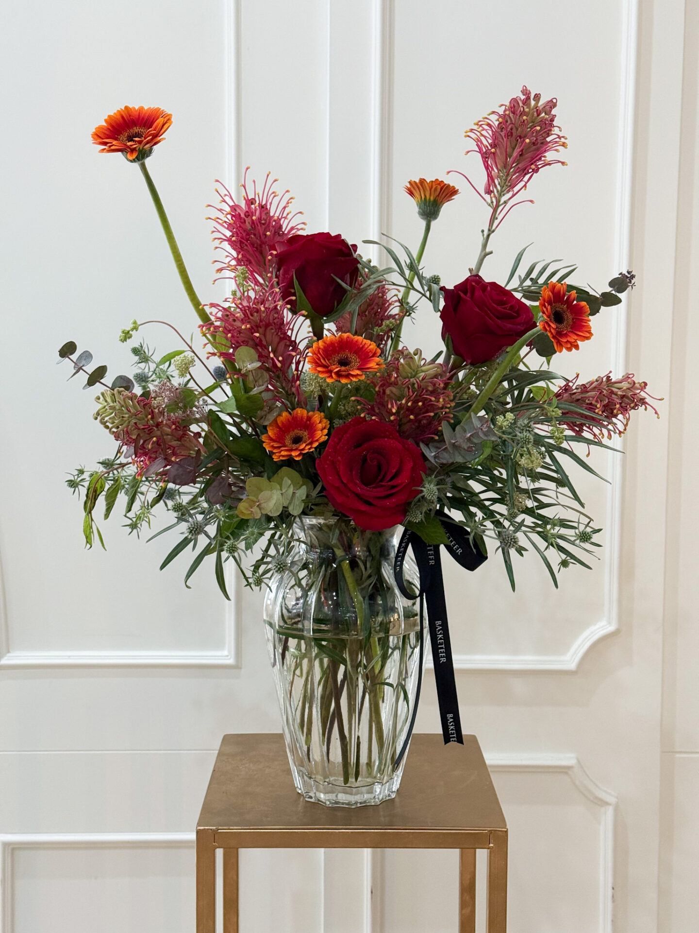 Modern vase arrangement with red roses, orange gerbera, and exotic red blooms in a clear glass vase. แจกันโมเดิร์น จัดด้วยกุหลาบแดง เยอบีร่าส้ม และดอกไม้สีแดงแปลกตา ในแจกันแก้วใส