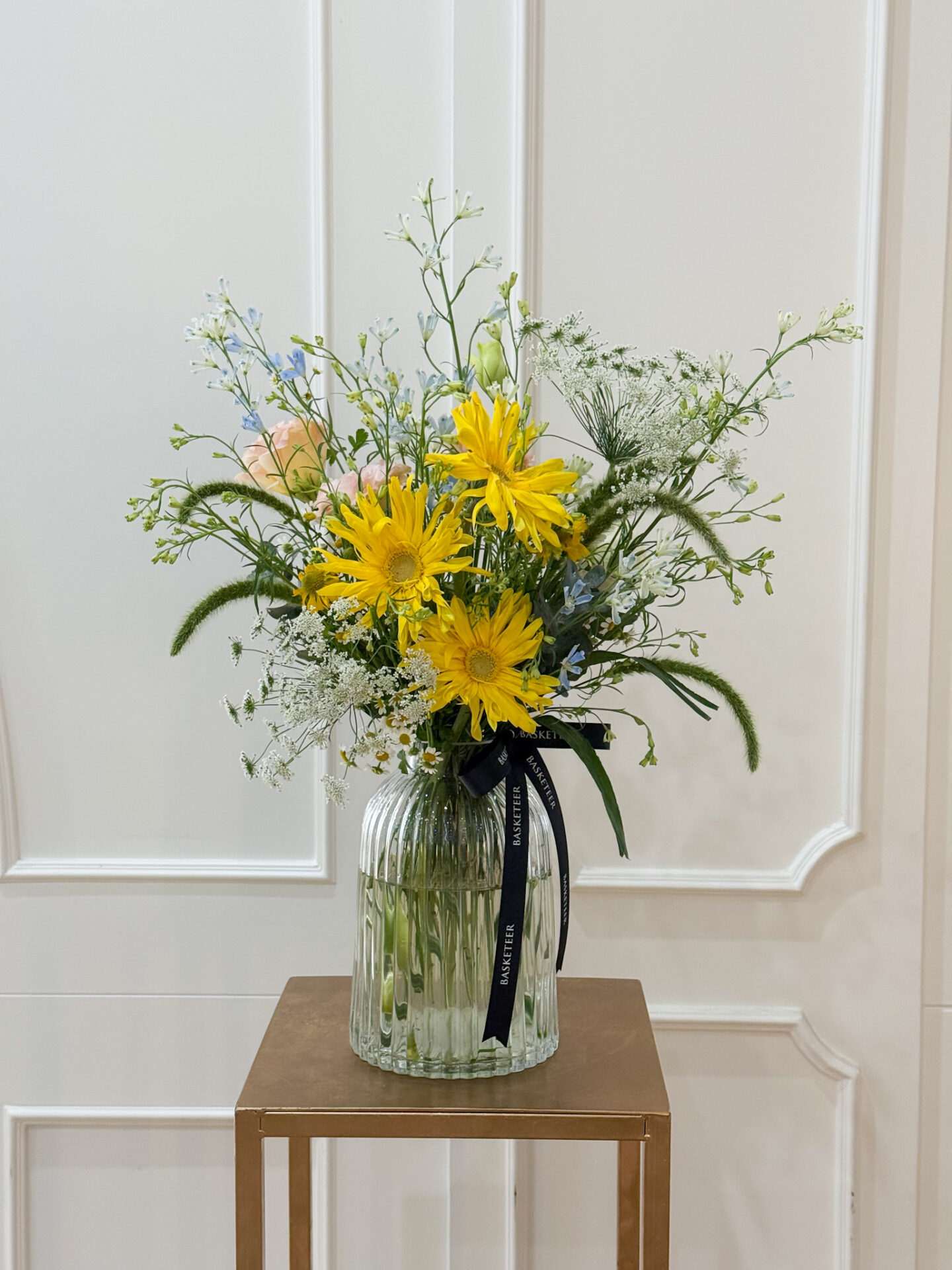 Tall glass vase arrangement with yellow gerbera, pastel flowers, and airy white fillers for a fresh, cheerful look.