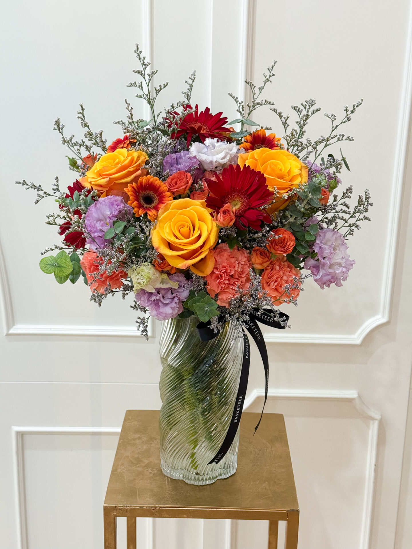 Tall glass vase arrangement with yellow roses, red gerbera, orange carnations, and purple blooms for a vibrant, elegant gift.