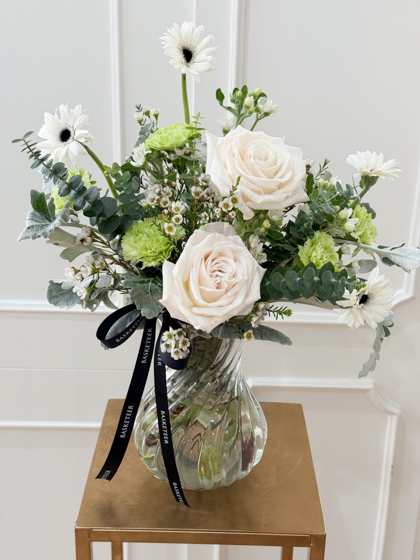 Glass vase with white roses, green carnations, white gerberas, and fresh greenery in a modern elegant design.