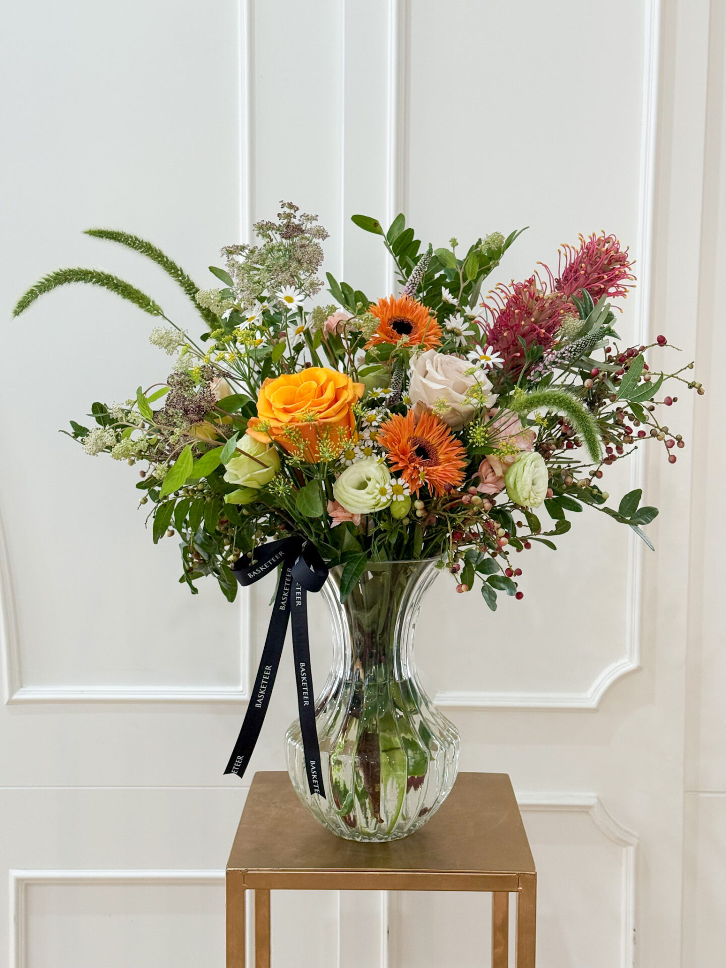 Glass vase arrangement with yellow roses, nude roses, orange gerberas, pink protea, and wild fillers for a vibrant elegant look.