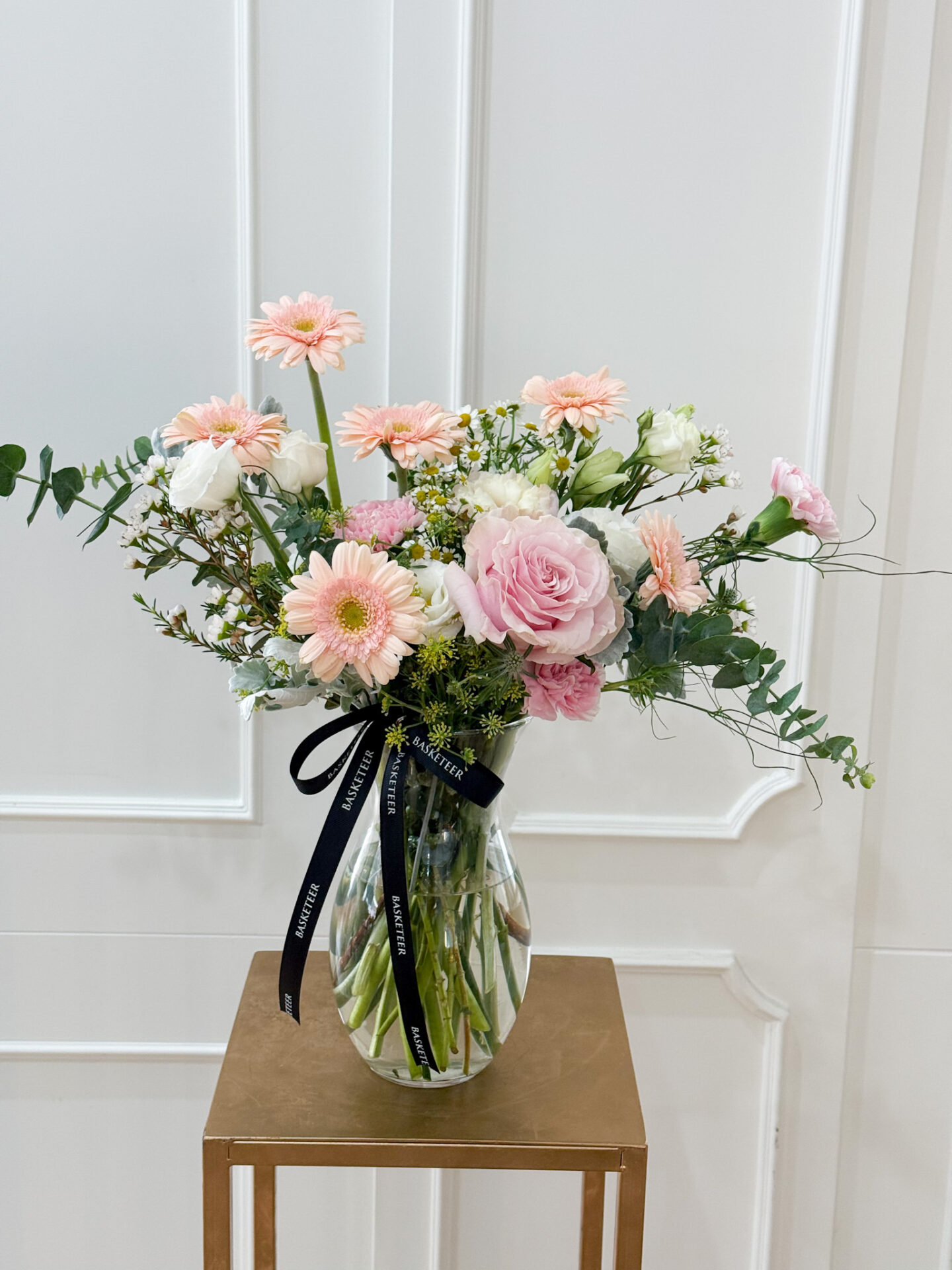 Glass vase with pastel pink roses, peach gerberas, white lisianthus, carnations, and fresh greenery for a soft romantic look.
