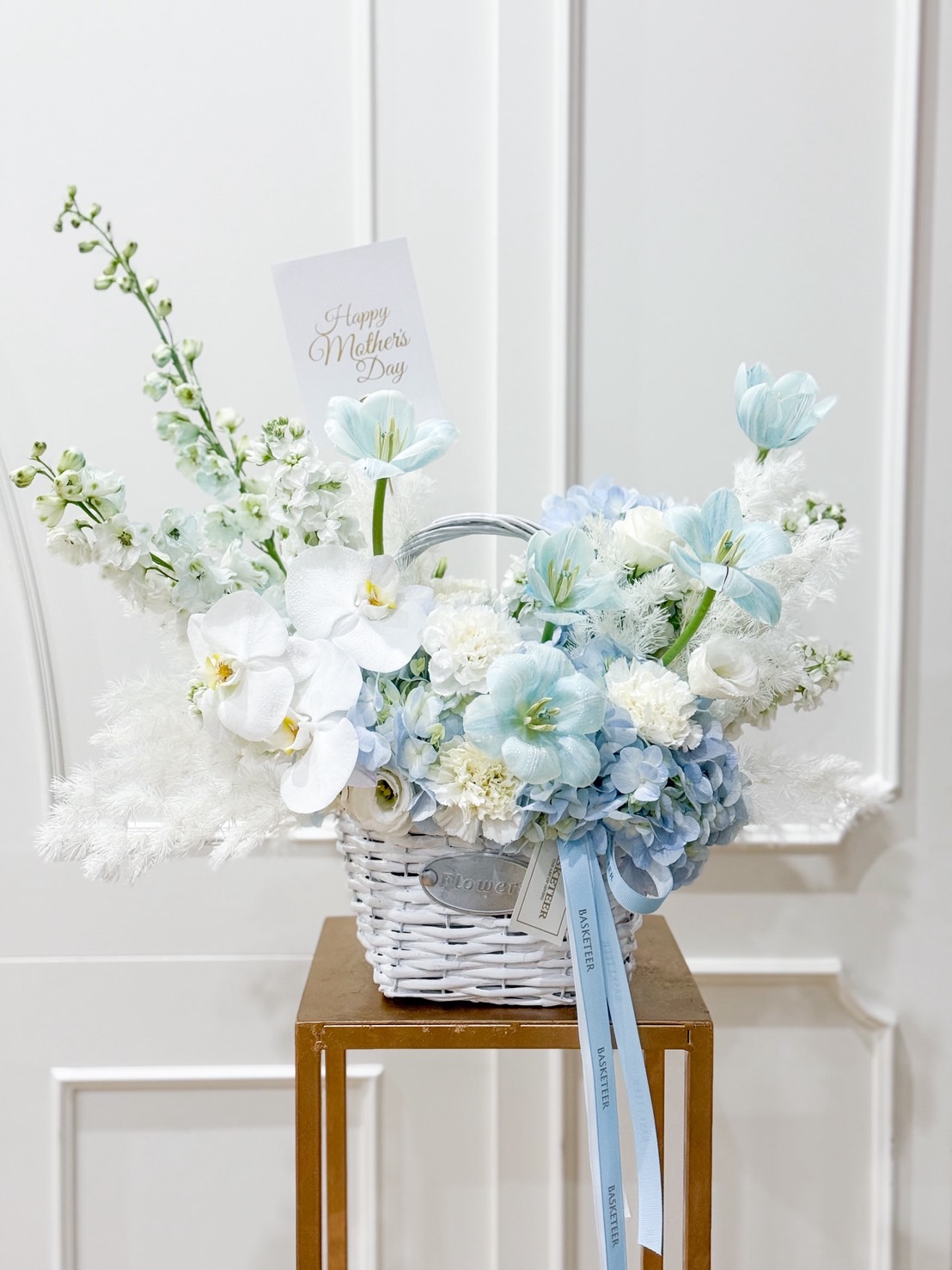 Luxury Mother’s Day flower basket in pastel blue and white, featuring tulips, orchids, hydrangeas, and carnations with “Happy Mother’s Day” card.