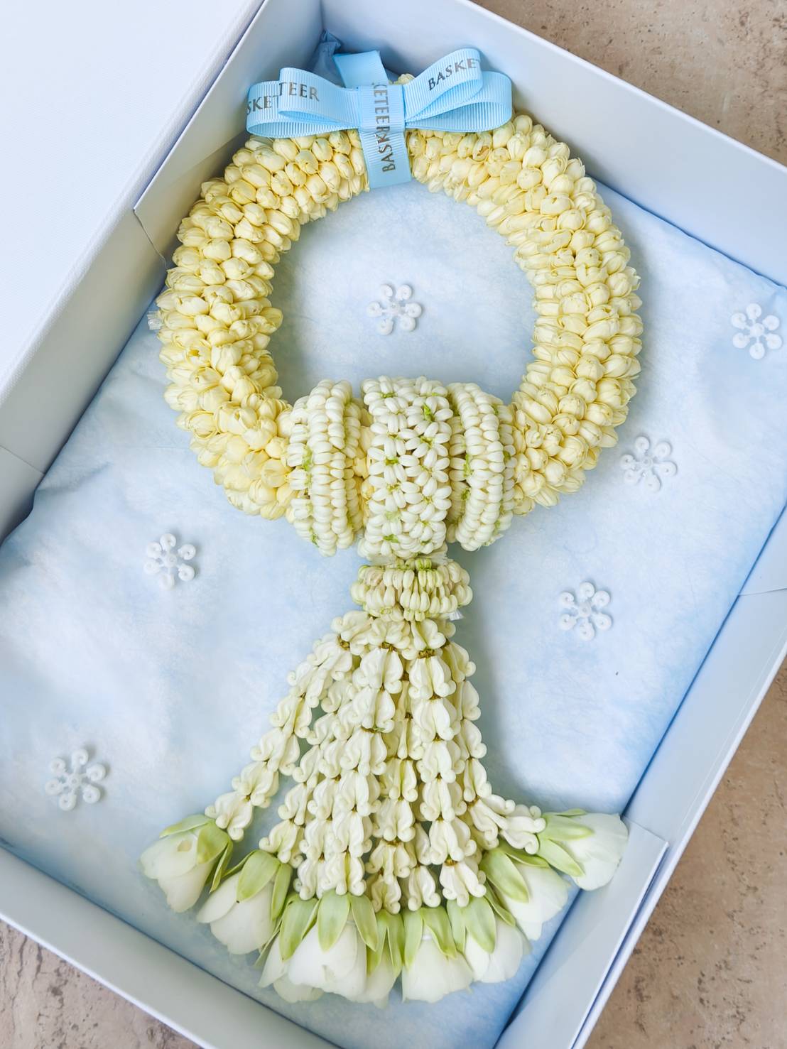 Premium cream jasmine garland with white rose buds, tied with pastel blue “Basketeer” ribbon, presented in an elegant gift box.