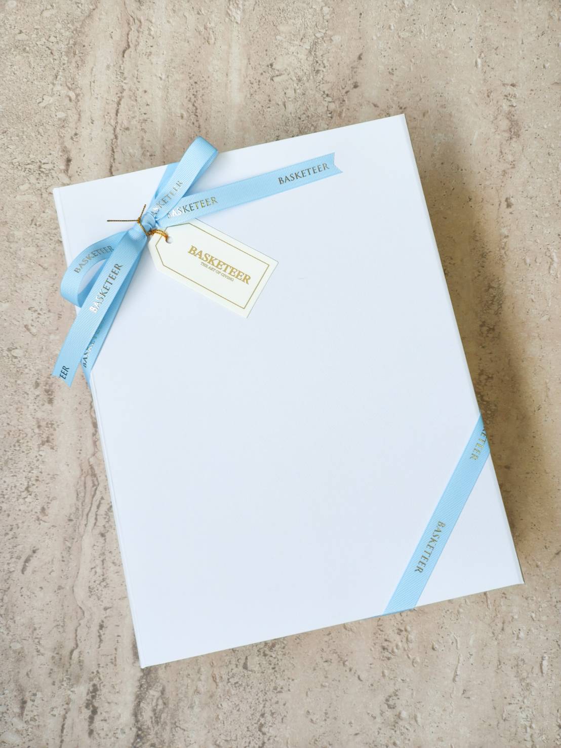Elegant white gift box tied with pastel blue “Basketeer” ribbon and gift tag, luxury packaging for premium presents.