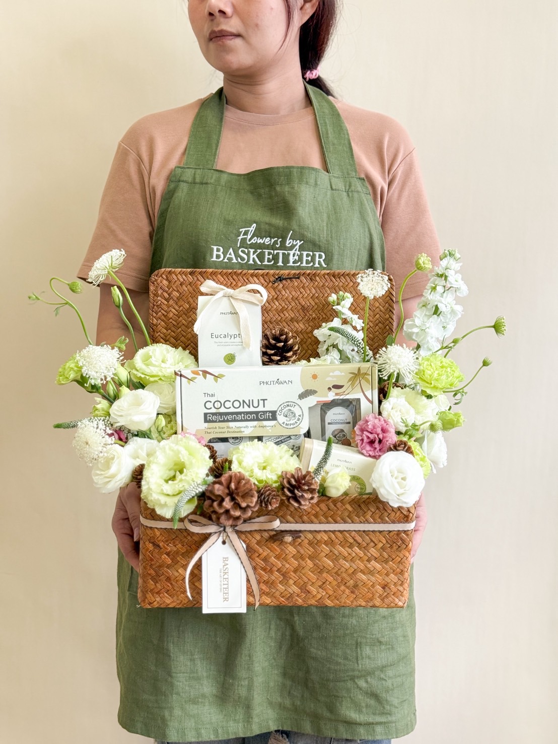 Person holding a woven spa hamper in white and green tones, featuring Thai coconut rejuvenation set, eucalyptus products, and fresh flowers.