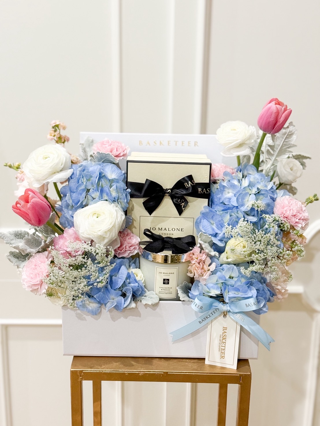 Elegant Jo Malone gift box surrounded by pastel pink, blue, and white flowers including tulips, hydrangeas, and ranunculus.