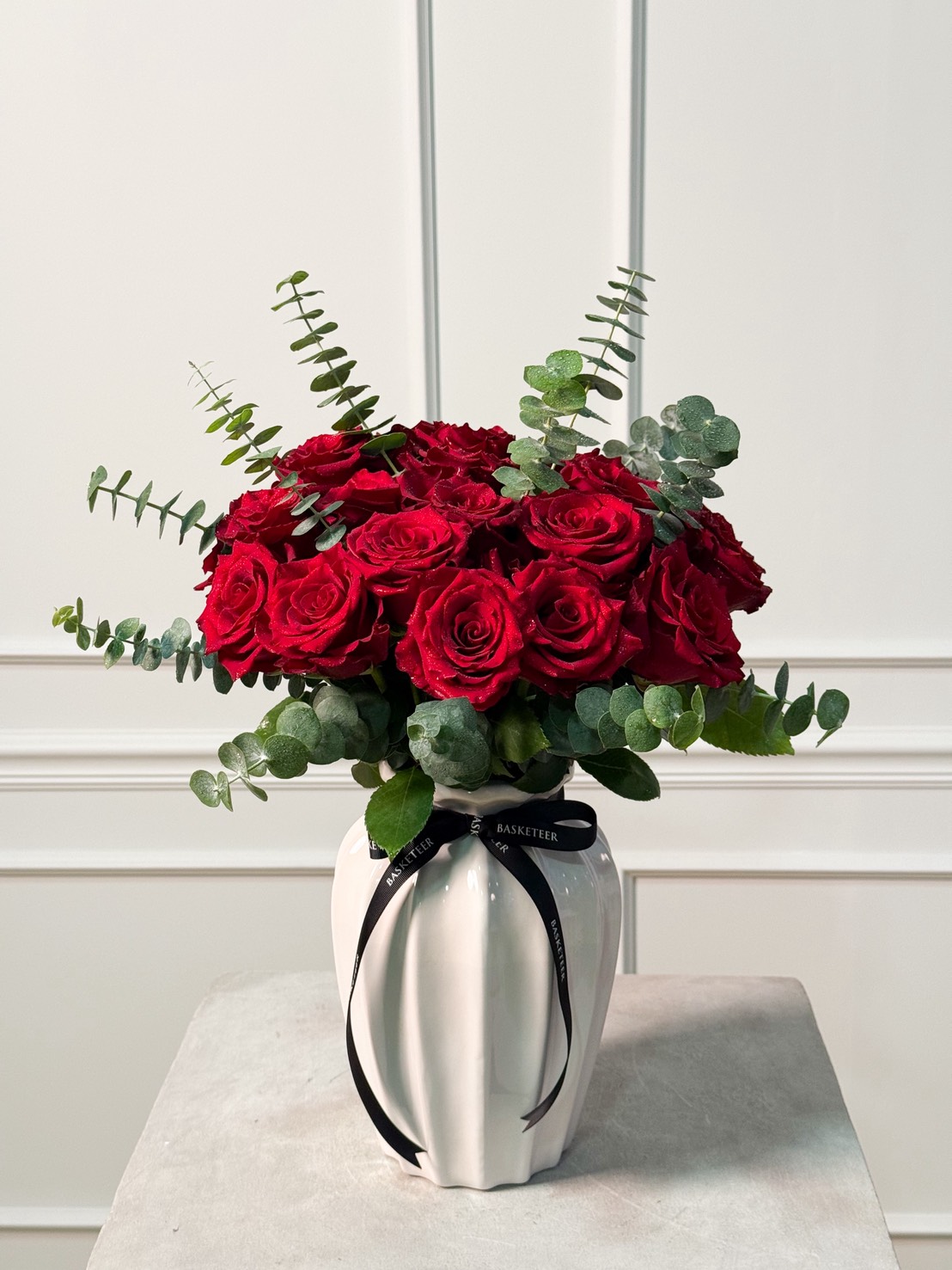 Elegant white ceramic vase filled with red roses and eucalyptus leaves, tied with a black Basketeer ribbon.