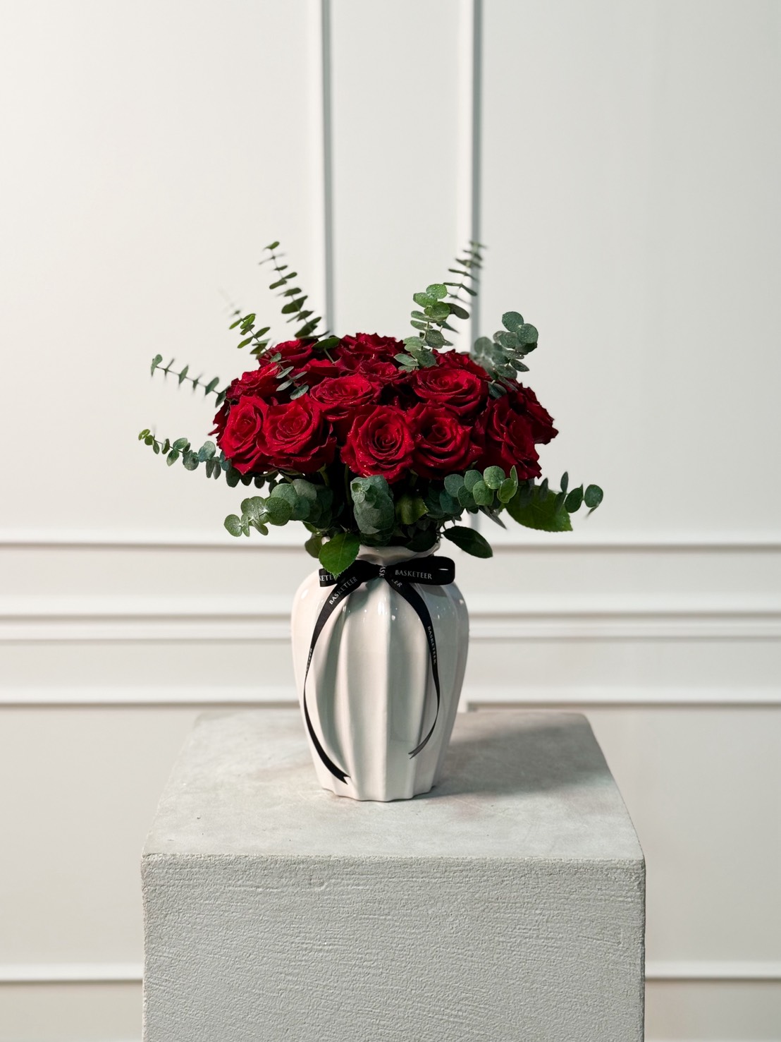 Front-view white ceramic vase filled with red roses and eucalyptus leaves, tied with a black Basketeer ribbon.
