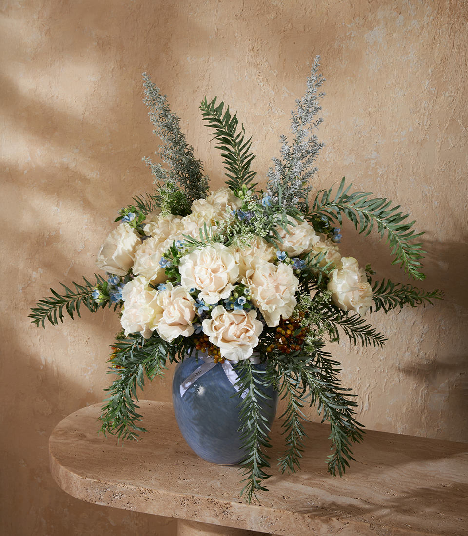 Elegant vase arrangement of cream carnations and roses, accented with blue fillers and lush greenery in a blue vase.