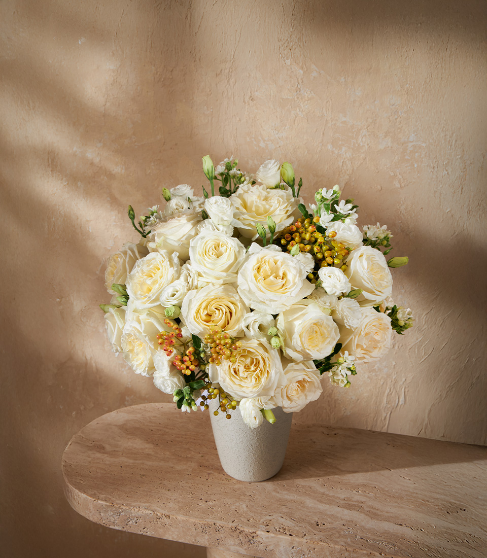 : Elegant vase of cream and white roses with delicate fillers and hypericum berries, arranged in a modern stone-textured vase.