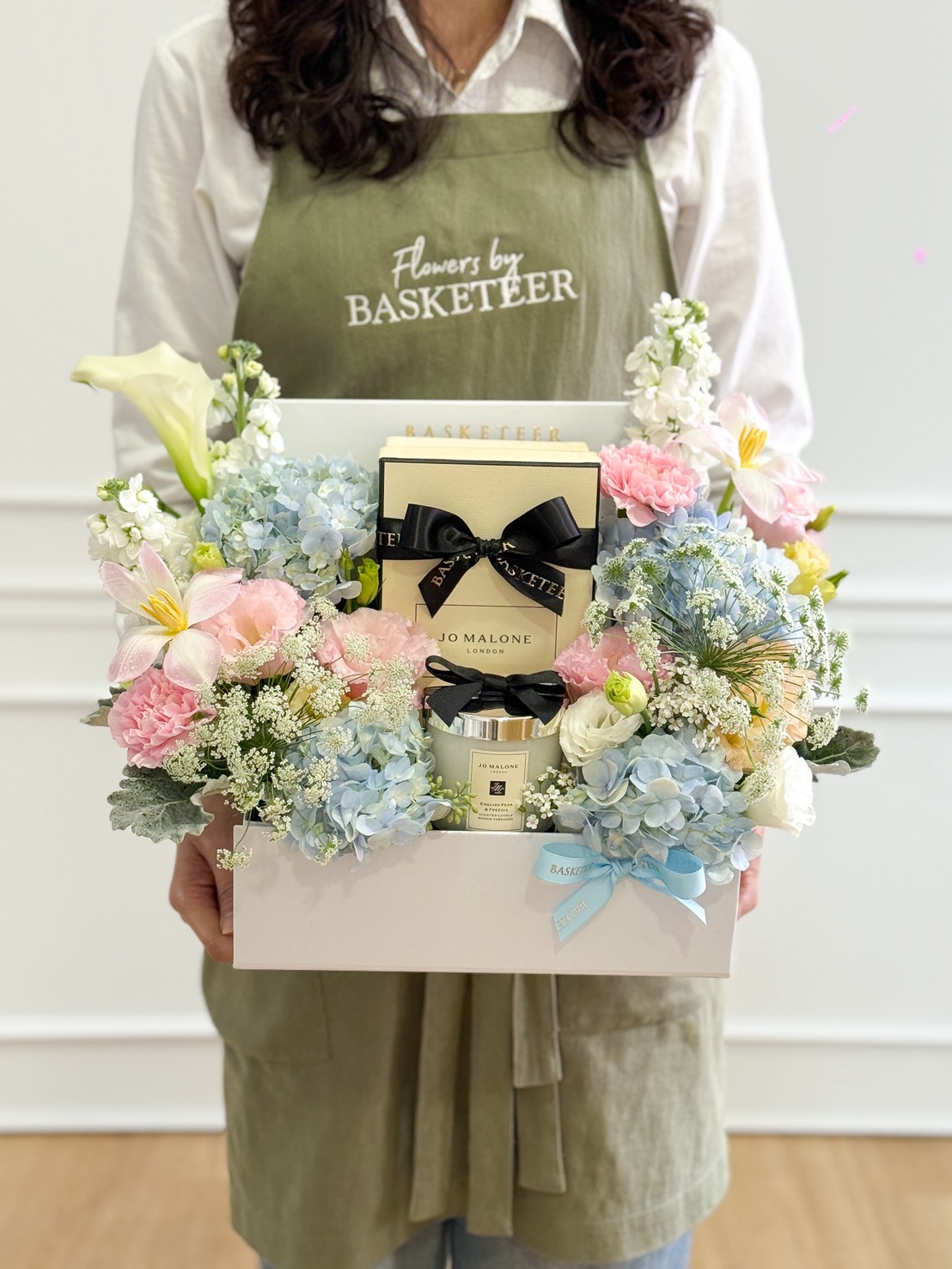 A person in a green apron holds a Jo Malone Candle Gift Box with Pastel Flowers, featuring a white box filled with flowers and luxury gift items, beautifully arranged for a charming floral gift presentation.