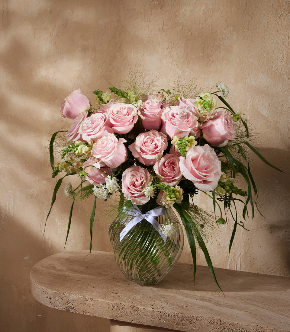Glass vase filled with soft pink roses, white fillers, and greenery, styled in a romantic floral arrangement.