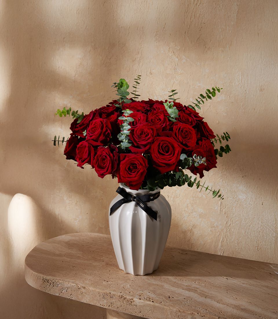 White vase filled with deep red roses and eucalyptus, styled with a black ribbon for a luxury floral arrangement.