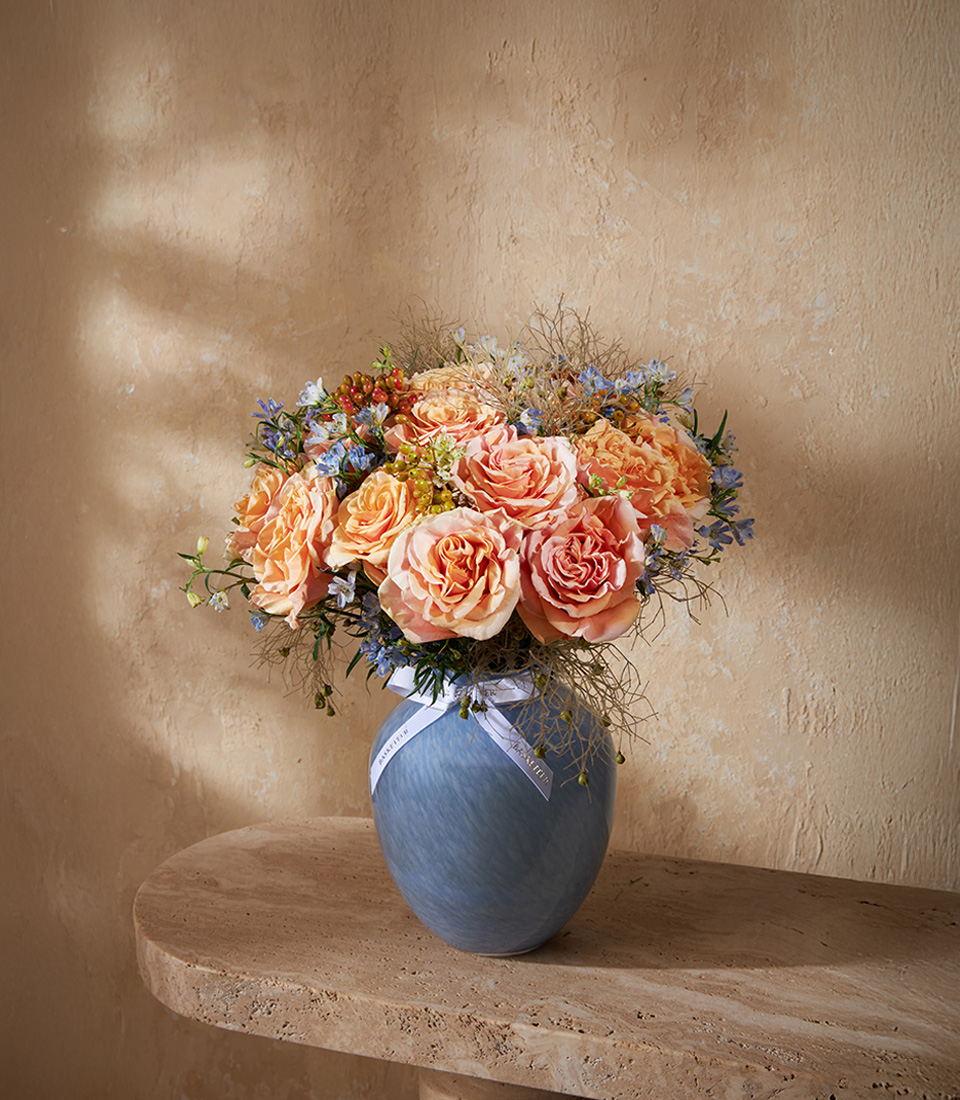 Elegant vase with peach-orange roses and delicate blue accents, arranged in a pastel blue ceramic vase for a luxury floral design.