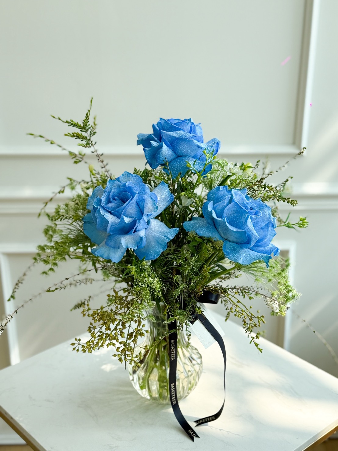 The Whisper Blue Elegance Vase Arrangement features three large blue roses in a clear glass vase, surrounded by lush green foliage and sprigs. A black ribbon accents the vase, all set on a white surface against a light-panelled wall.