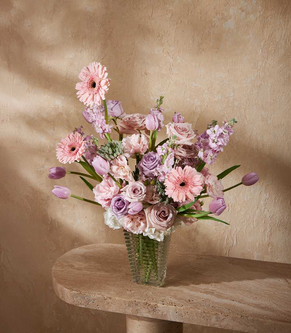 Elegant flower arrangement with pink and purple roses, tulips, gerbera daisies, and hydrangeas in a tall glass vase.