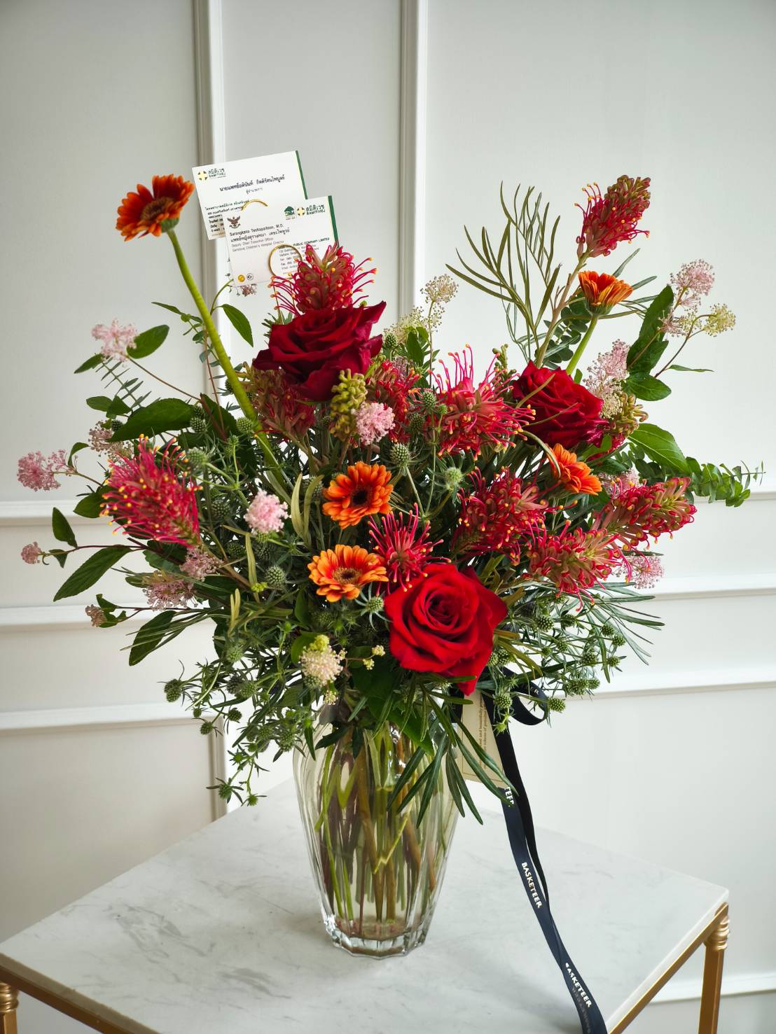 Vibrant vase arrangement with red roses, orange gerberas, tropical blooms, and greenery in a clear glass vase