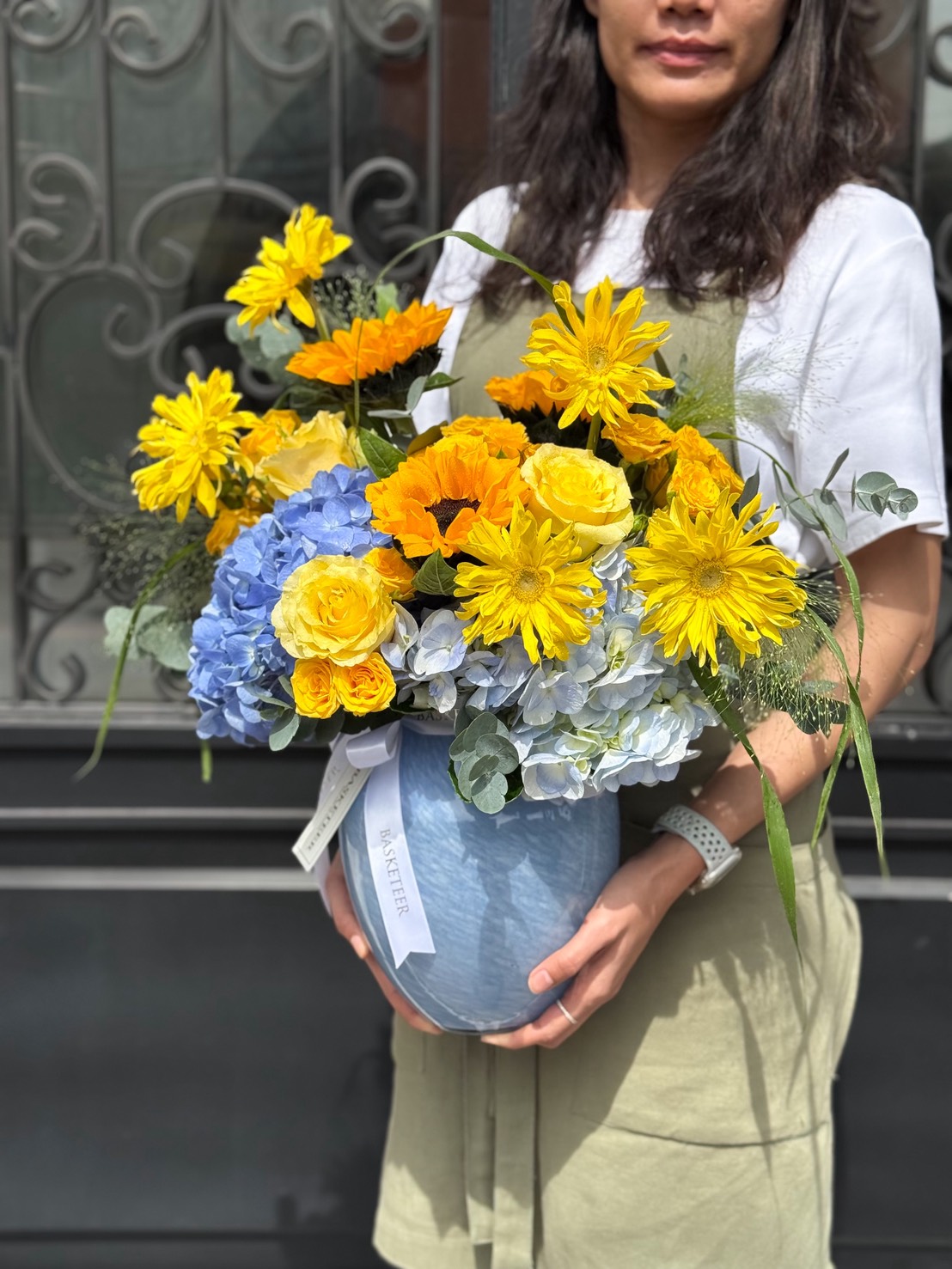 A woman wearing a white shirt and beige apron holds the Golden Horizon Vase, filled with yellow and orange flowers, blue hydrangeas, and greenery, standing in front of a decorative black metal gate.