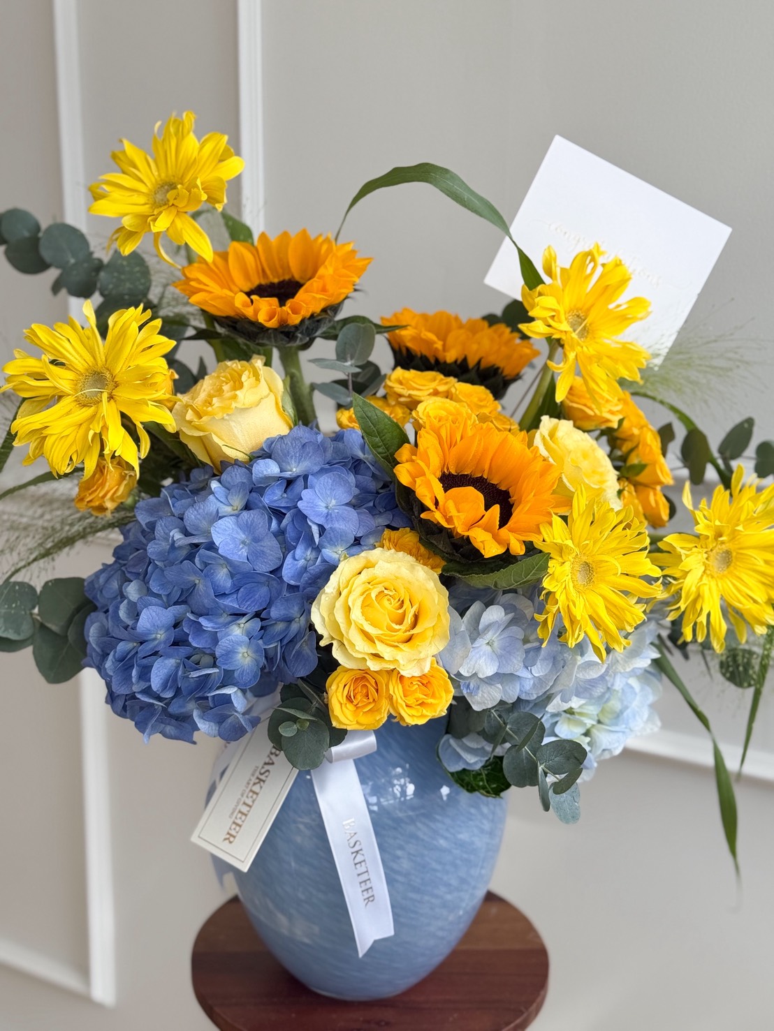 The Golden Horizon Vase holds a bright bouquet of yellow sunflowers, yellow roses, blue hydrangeas, and green foliage, with a white card and an “EASTER” ribbon. The arrangement sits on a small wooden table.
