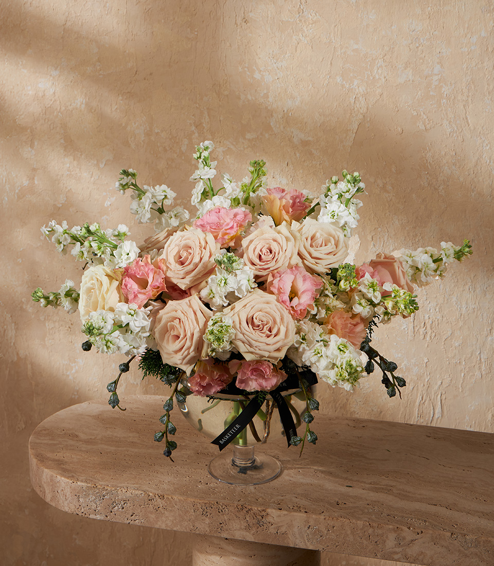 Pastel flower arrangement with cream and pink roses, white stock, and soft greenery in a compote vase – luxury flower delivery Bangkok.