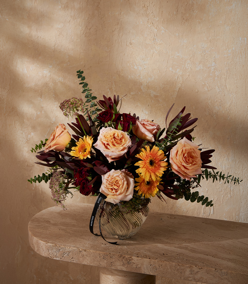 Elegant vase with peach roses, yellow gerberas, and burgundy accents, arranged with greenery in a stylish round glass vase.
