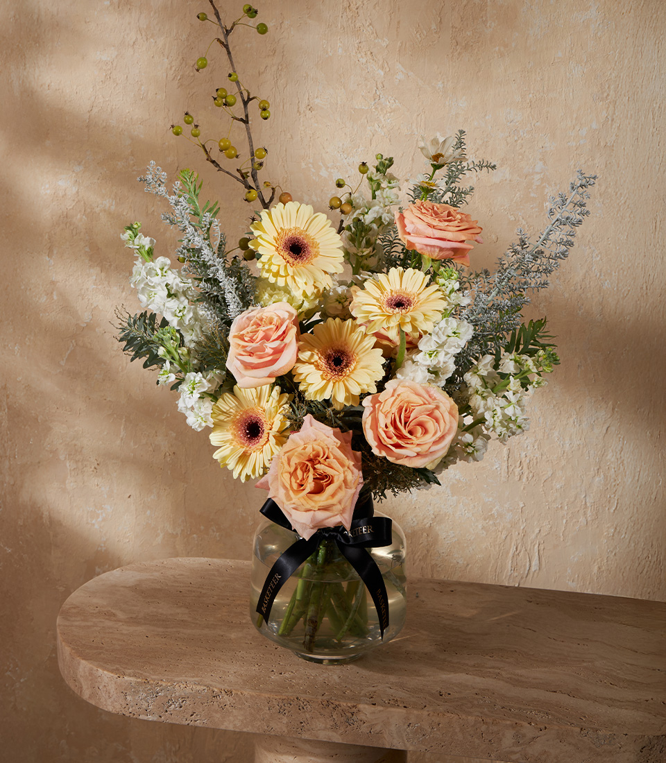 Elegant vase with peach roses, yellow gerberas, white stock, and greenery, styled with natural soft textures for a warm display.