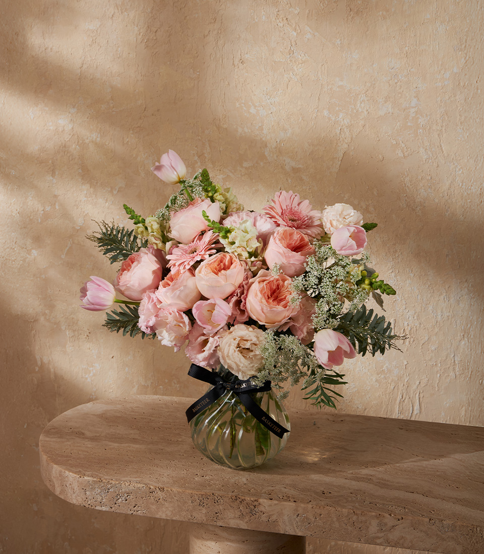 Elegant vase with pink garden roses, tulips, and pastel blooms, arranged with greenery in a round glass vase.