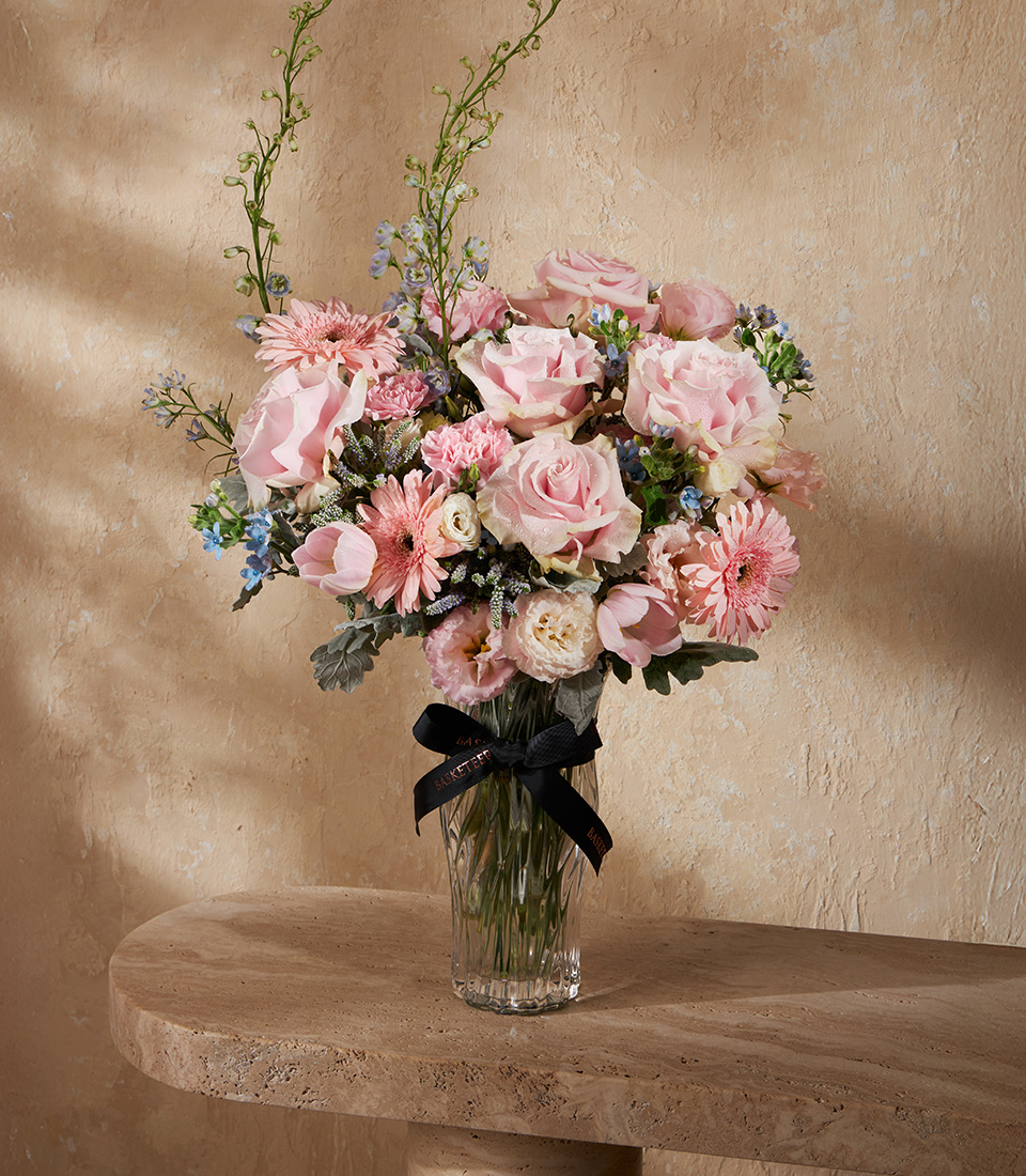 Elegant vase arrangement of pink roses, gerberas, tulips, and carnations, accented with delicate blue blooms and soft greenery.