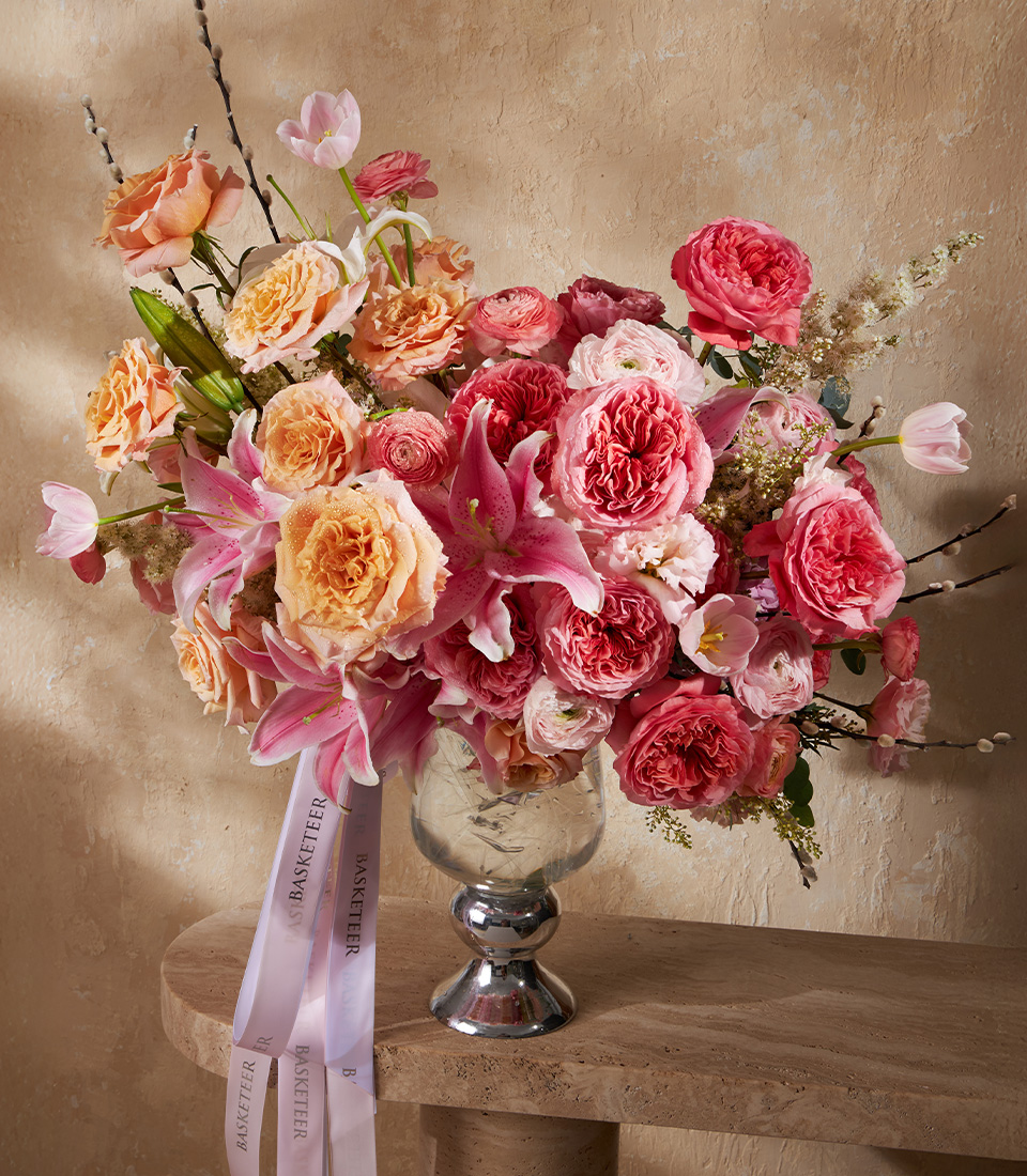 Luxurious tall vase with pink and peach roses, lilies, and ranunculus, styled with greenery and soft blooms for a grand arrangement.
