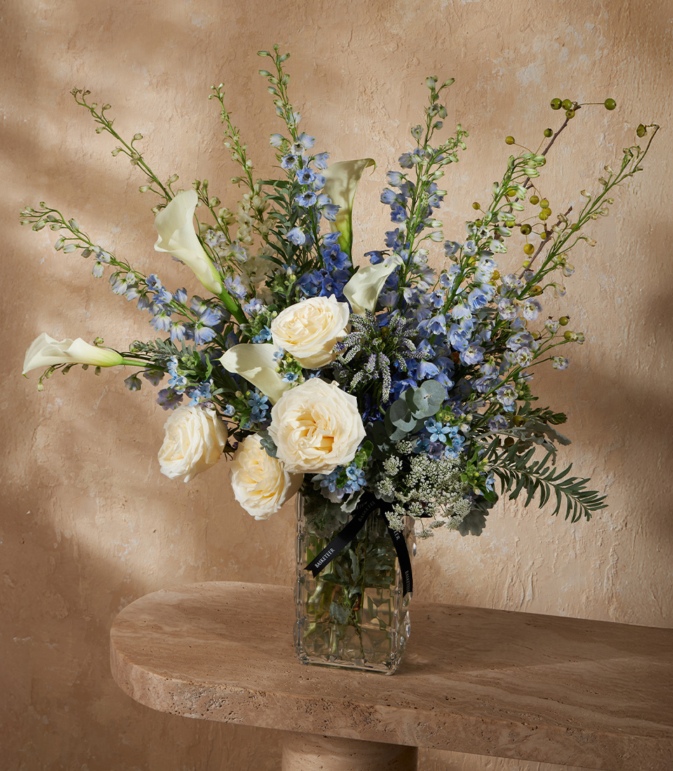 Elegant tall vase with white roses, calla lilies, and blue delphiniums, accented with lush greenery for a refined floral arrangement.