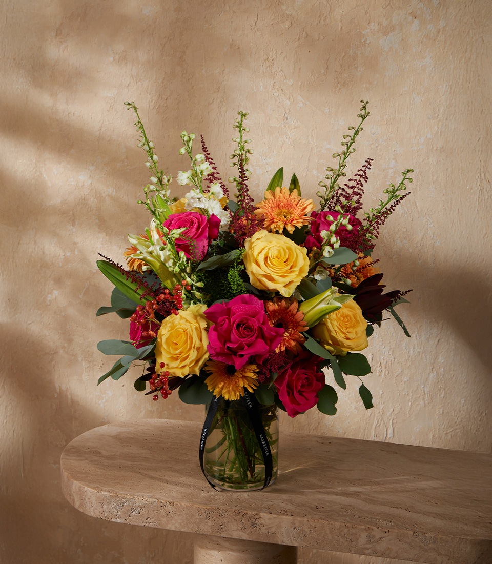 Bright vase arrangement of yellow roses, hot pink roses, orange gerberas, and lilies, accented with lush greenery and astilbe.