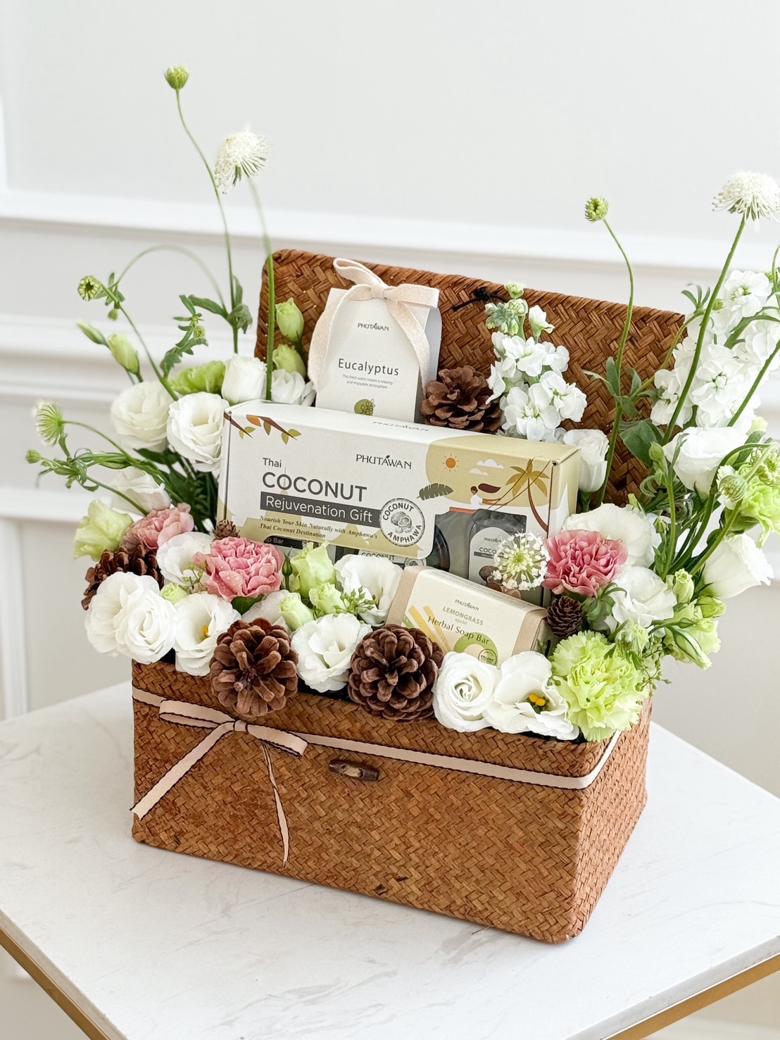 Natural woven basket with Phutawan spa gift set, decorated with white lisianthus, carnations, hydrangea accents, and pinecones.