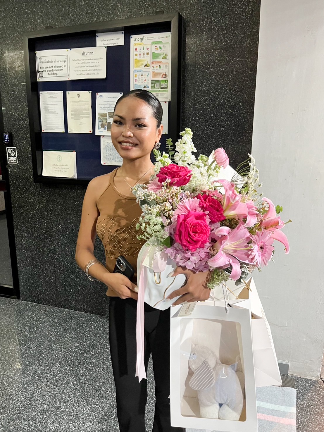 Woman holding a pink flower bouquet with roses, lilies, gerberas, and hydrangeas, paired with a gift bag containing a plush toy.