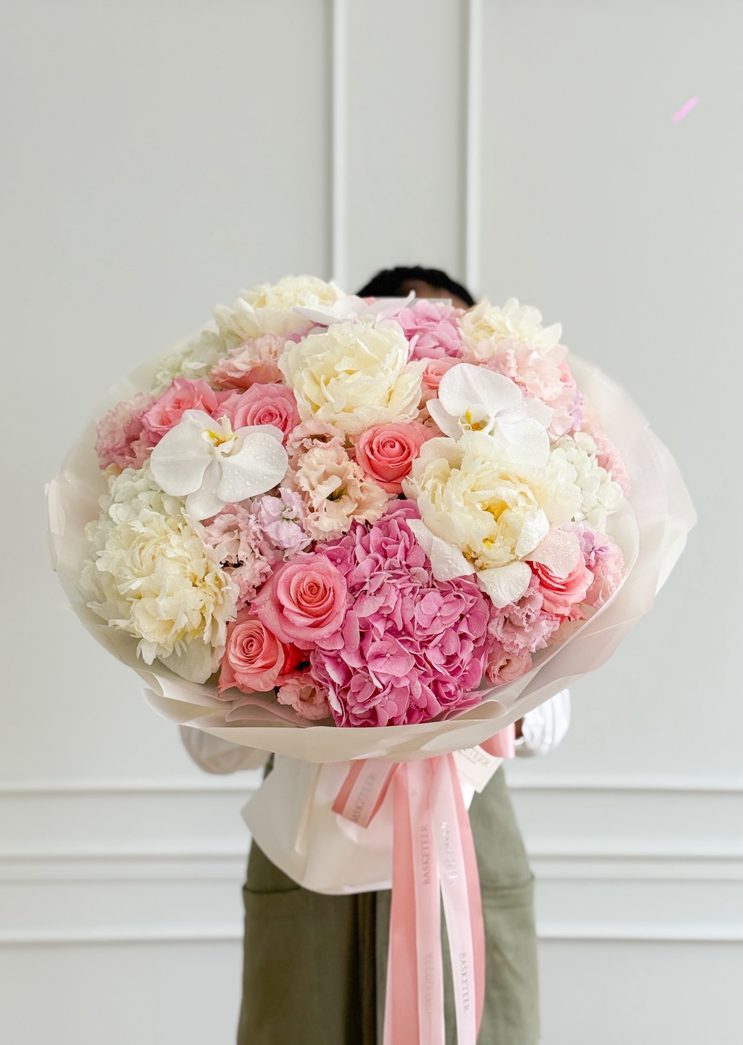 A person holding a Premium Mixed Peony Bouquet of pink and white flowers, including roses, orchids, and peonies, wrapped in light paper with a pink ribbon against a plain white background.