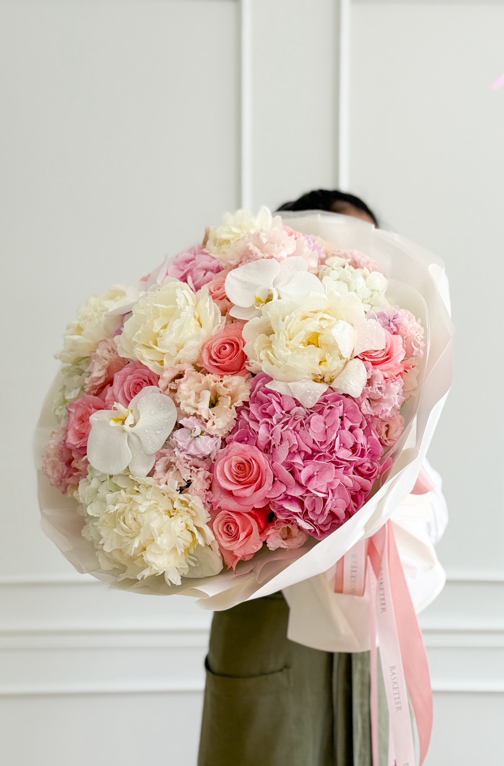 A person holds a Premium Mixed Peony Bouquet featuring pink, white, and cream flowers—roses, hydrangeas, and peonies—wrapped in white paper with a pink ribbon against a plain white wall background.