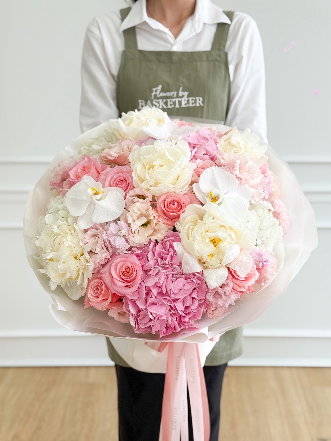 A person wearing a green apron holds a Premium Mixed Peony Bouquet—a large arrangement of pink and white flowers, including roses, hydrangeas, peonies, and white orchids—tied elegantly with a pink ribbon.